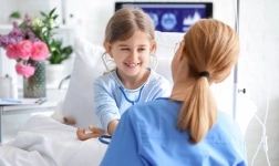 A young girl sits on a hospital bed smiling at a doctor during a checkup, with flowers and medical equipment in the background.